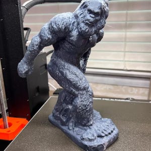 May include: A 3D-printed figurine of Bigfoot in a gray color. The sculpture depicts the legendary creature in a walking pose, with detailed fur texture and a large footprint base. The figure is on a 3D printer bed.