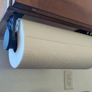 May include: A white paper towel roll is mounted under a wooden cabinet. The roll is held in place by a black holder. The paper towels have a textured surface and are ready for use in the kitchen.