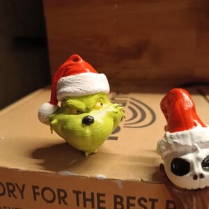May include: A green Grinch figure wearing a red and white Santa hat. Next to it is a white skull figure with a red and white Santa hat. Both figures are on a cardboard surface.