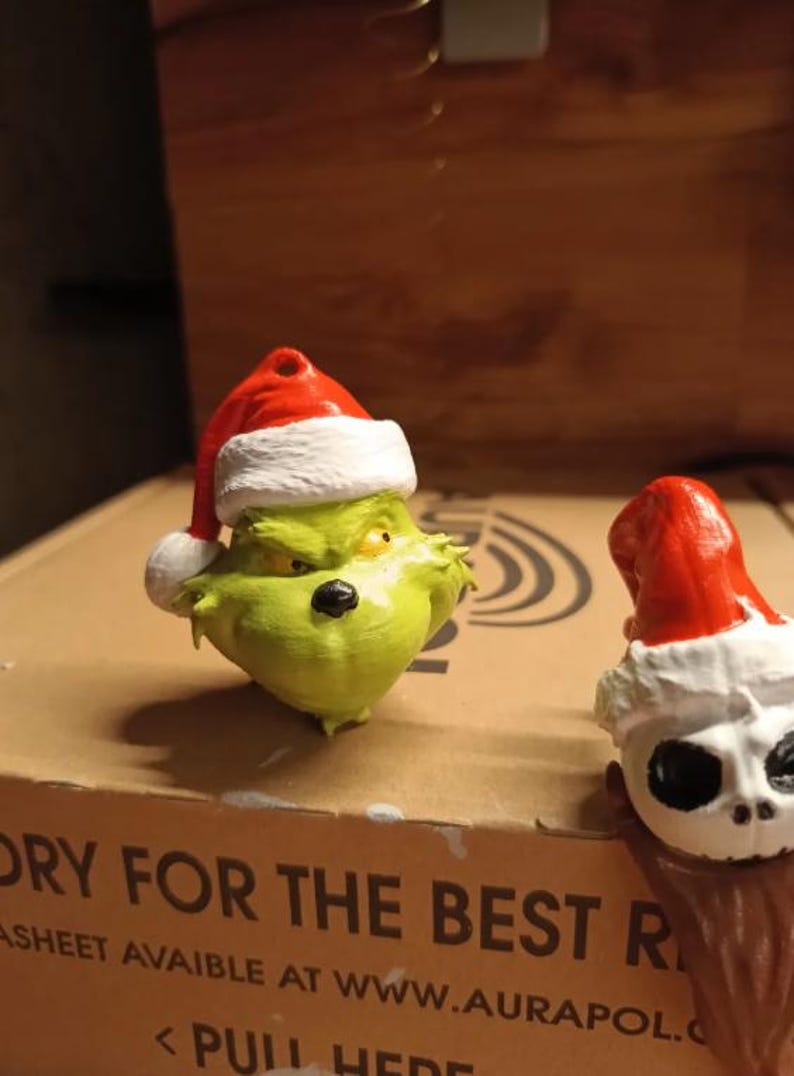 May include: A green Grinch figure wearing a red and white Santa hat. Next to it is a white skull figure with a red and white Santa hat. Both figures are on a cardboard surface.
