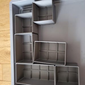 Peut inclure: Plusieurs contenants carrés gris de différentes tailles sont disposés dans un grand plateau rectangulaire gris. Les contenants présentent un motif quadrillé sur le fond et sont faits d'un matériau robuste.