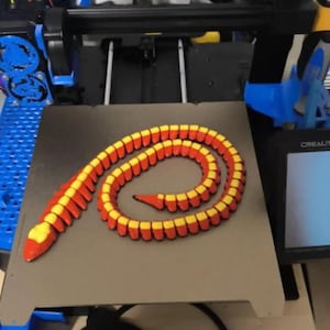 Puede incluir: Una serpiente articulada impresa en 3D, roja y amarilla, enrollada en una cama de impresora. La impresora 3D es azul y negra, con una pantalla digital. Se ven componentes adicionales, como una tarjeta de memoria y una pieza circular negra.