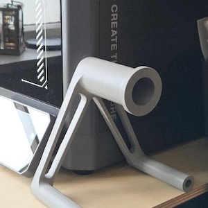 May include: A light grey, 3D-printed object with a cylindrical top and two angled legs. The object is designed to hold something, with a hole at the top and at the end of each leg. The text "CREATE T" is visible in the background.