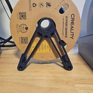 Puede incluir: Un soporte negro para bobina de filamento de impresora 3D que sostiene una bobina de cart&oacute;n marr&oacute;n de filamento azul. La bobina est&aacute; etiquetada como "CREALITY HYPER SERIES 3D PRINTER FILAMENT". El soporte est&aacute; sobre una superficie de madera.