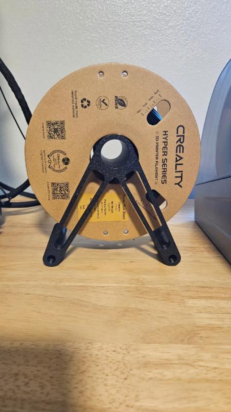 Puede incluir: Un soporte negro para bobina de filamento de impresora 3D que sostiene una bobina de cart&oacute;n marr&oacute;n de filamento azul. La bobina est&aacute; etiquetada como "CREALITY HYPER SERIES 3D PRINTER FILAMENT". El soporte est&aacute; sobre una superficie de madera.
