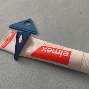 Puede incluir: Un tubo de pasta de dientes "elmex" con un diseño rojo y blanco, acompañado de dos exprimidores de tubo de pasta de dientes triangulares de plástico azul. Un exprimidor está unido al tubo, el otro está separado.