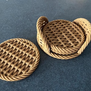 Op de afbeelding: Set van twee geweven rotan onderzetters. Eén onderzetter is los, de andere drie zijn gestapeld met een geweven handvat. De onderzetters zijn lichtbruin van kleur en hebben een ronde vorm. Het weefpatroon is zichtbaar. Diameter ongeveer 10 cm.