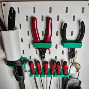 May include: A white pegboard with various tools organized on it. Tools include pliers with red handles, black-handled pliers, files with red handles, and a digital caliper. A white cup holds pens.