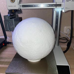 May include: A white, textured sphere sits on a 3D printer bed. The printer, a Bambu Lab model, has a white frame and a moving print head. The sphere appears to be a model or prototype, possibly for a craft or design project.