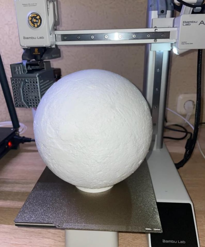 May include: A white, textured sphere sits on a 3D printer bed. The printer, a Bambu Lab model, has a white frame and a moving print head. The sphere appears to be a model or prototype, possibly for a craft or design project.