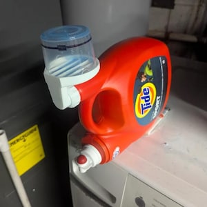 May include: A red Tide Sport laundry detergent bottle with a blue and white measuring cup attached. The bottle is resting on a white washing machine. The detergent bottle has a yellow and blue label.