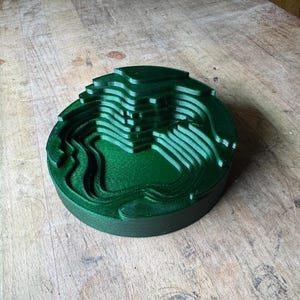 May include: A dark green, layered, circular object with a topographical design. The object has a series of concentric rings, creating a 3D effect. The object is sitting on a wooden surface.