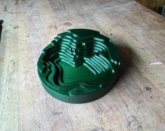 Può includere: Un oggetto circolare verde scuro, a strati, con un design topografico. L'oggetto ha una serie di anelli concentrici, creando un effetto 3D. L'oggetto è appoggiato su una superficie di legno.