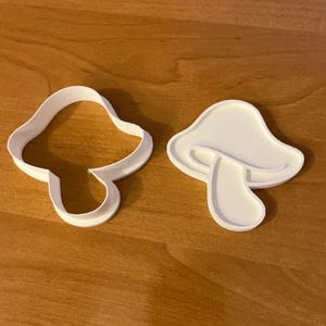 May include: Two white mushroom-shaped cookie cutters. One is an outline, and the other is a solid shape. The cutters are made of plastic and sit on a wooden surface. Ideal for baking.