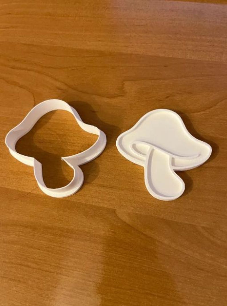 May include: Two white mushroom-shaped cookie cutters. One is an outline, and the other is a solid shape. The cutters are made of plastic and sit on a wooden surface. Ideal for baking.