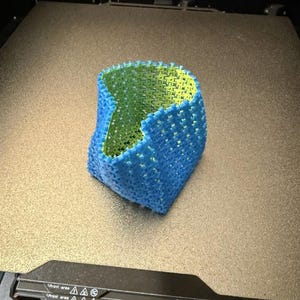 May include: A 3D-printed object with a blue exterior and a green interior, showcasing a woven or lattice-like pattern. The object is on a Prusa XL 3D printer bed, highlighting its creation process.