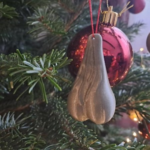 Könnte beinhalten: Ein neuartiger Weihnachtsbaumschmuck in Form von Hoden, in hellgrauer Farbe. Der Schmuck hängt an einer roten Schnur, mit einer roten und goldenen Weihnachtskugel im Hintergrund, umgeben von grünen Tannennadeln.