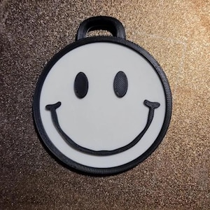 May include: A round, white smiley face pendant with black eyes and a curved mouth. The pendant has a black border and a loop at the top for hanging. The design is simple and cheerful, suitable for a keychain or charm.