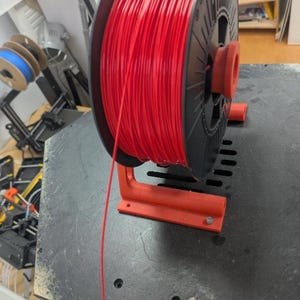 Puede incluir: Una bobina de filamento rojo para impresi&oacute;n 3D, montada en un soporte rojo. La bobina negra contiene una gran cantidad de filamento rojo. El soporte est&aacute; fijado a una superficie oscura. Se ven bobinas adicionales en el fondo.