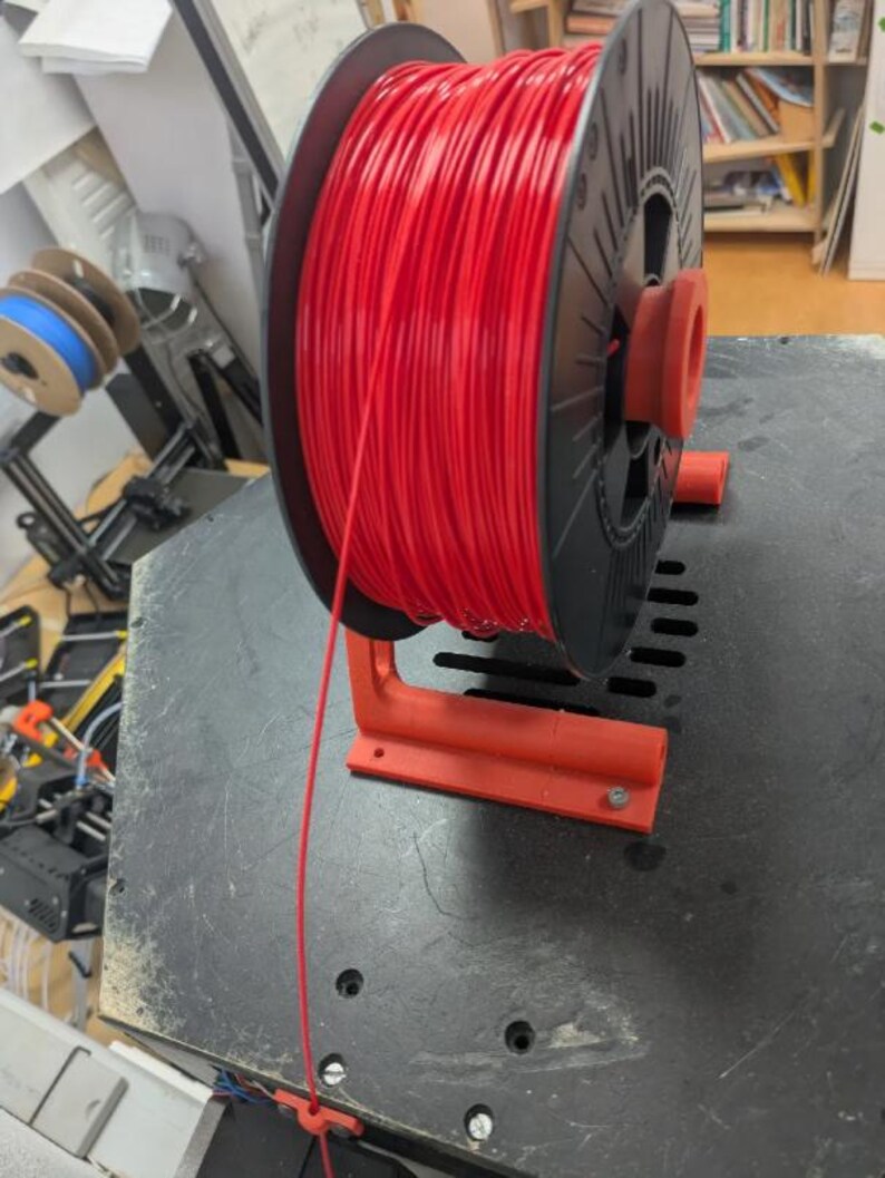 Puede incluir: Una bobina de filamento rojo para impresi&oacute;n 3D, montada en un soporte rojo. La bobina negra contiene una gran cantidad de filamento rojo. El soporte est&aacute; fijado a una superficie oscura. Se ven bobinas adicionales en el fondo.