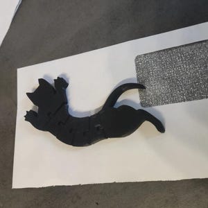May include: A black cat-shaped puzzle is assembled on a white envelope. The cat is in a playful pose, with its paws extended. The puzzle is made of several pieces, and the cat's form is clearly defined.