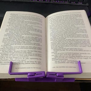 May include: Open book with a purple book holder. The book is titled "THE HITCHHIKER'S GUIDE TO THE GALAXY" and has text on the pages. The book holder is made of plastic and is designed to hold the book open.