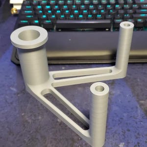 Puede incluir: Un soporte gris claro, impreso en 3D, con un dise&ntilde;o &uacute;nico. Cuenta con una gran abertura circular en la parte superior, dos soportes cil&iacute;ndricos y una base triangular con recortes. El objeto es probablemente un componente para un ensamblaje m&aacute;s grande.