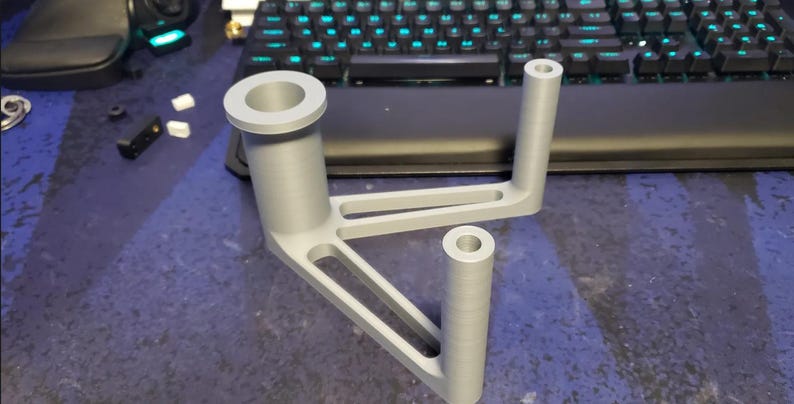 Puede incluir: Un soporte gris claro, impreso en 3D, con un dise&ntilde;o &uacute;nico. Cuenta con una gran abertura circular en la parte superior, dos soportes cil&iacute;ndricos y una base triangular con recortes. El objeto es probablemente un componente para un ensamblaje m&aacute;s grande.