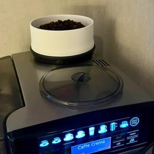 May include: A silver and black espresso machine with a clear lid and a white container filled with coffee beans. The machine's display shows various coffee icons and the text "Caffe Crema". The "start stop" button is visible.