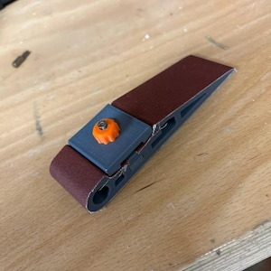 Puede incluir: Una herramienta de lijado manual con una banda abrasiva granate. La herramienta tiene un marco gris con una perilla de ajuste naranja. La herramienta está diseñada para lijar madera y otros materiales. Se muestra sobre una superficie de madera.