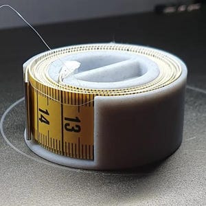 以下が含まれることがあります： 黒い目盛りの付いた黄色い巻き尺で、13インチと14インチの数字が表示されています。薄いグレーのホルダーに収められています。テープは部分的にほどかれ、細い白い糸が出ています。背景はぼやけています。