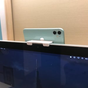 Puede incluir: Un teléfono inteligente verde claro está montado en un clip blanco, sujeto a la parte superior de un monitor de computadora. El monitor muestra una pantalla azul oscuro. La configuración está contra una pared beige claro.