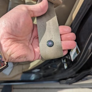 Pode incluir: Cinto de segurança bege com um botão preto com um design roxo. O cinto de segurança é segurado por uma mão, com a fivela visível na parte superior. O interior do carro é visível ao fundo.