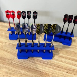 Peut inclure: Ensemble de fléchettes avec des plumes de différentes couleurs, dont le rouge, le noir et des motifs, présentées dans des supports bleus. Les fléchettes sont disposées sur une surface en bois, présentant une variété de styles.