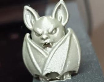 Könnte beinhalten: Silberfarbene, 3D-gedruckte Fledermausfigur. Die Fledermaus hat ein detailliertes Gesicht mit spitzen Ohren, einem grimmigen Ausdruck und ausgebreiteten Flügeln. Die Figur ist mit einer strukturierten Oberfläche versehen und steht auf einer flachen Basis.