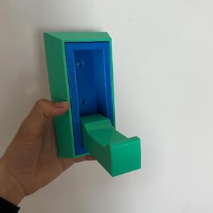 May include: A wall-mounted, two-tone hook in shades of green and blue. The hook is rectangular with a curved arm extending outward. The interior is a contrasting blue, and the exterior is a vibrant green. The hook is designed for hanging items.