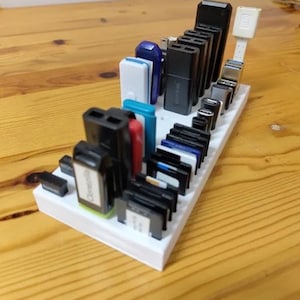 Puede incluir: Un organizador rectangular blanco que contiene varias unidades USB y adaptadores negros, azules, rojos y blancos. Los artículos están dispuestos en filas y columnas, mostrando una variedad de formas y tamaños. El organizador está sobre una superficie de madera marrón claro.