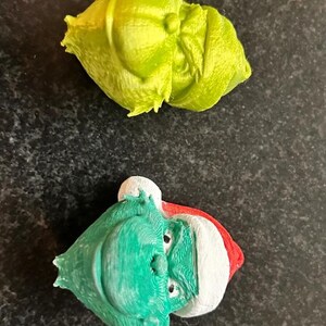 May include: Two 3D-printed Christmas ornaments. One is a lime green, abstract shape. The other is a green face with a white and red hat. The ornaments are on a dark, speckled surface.