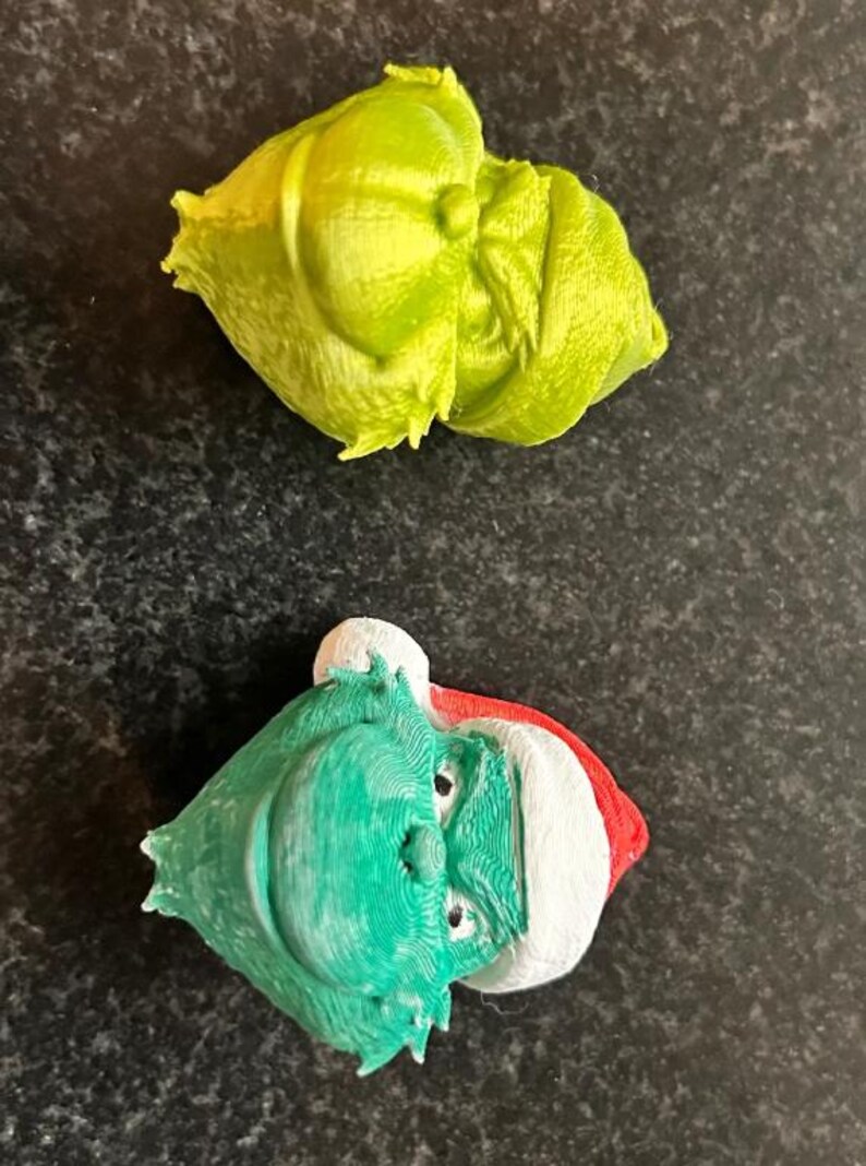 May include: Two 3D-printed Christmas ornaments. One is a lime green, abstract shape. The other is a green face with a white and red hat. The ornaments are on a dark, speckled surface.