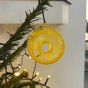 May include: A yellow, circular Christmas ornament with a woven design, hanging from a green pine branch. The ornament is illuminated by small, warm white lights, creating a festive holiday scene.