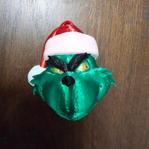 May include: A green Grinch head ornament with a Santa hat. The Grinch has a black nose, black eyebrows, and yellow eyes with red pupils. The Santa hat is red with a white trim.