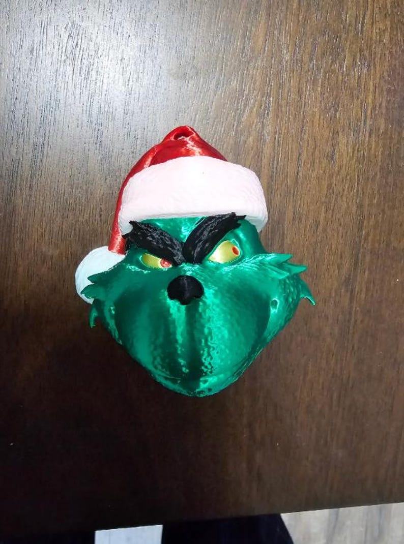 May include: A green Grinch head ornament with a Santa hat. The Grinch has a black nose, black eyebrows, and yellow eyes with red pupils. The Santa hat is red with a white trim.