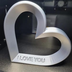 Può includere: Una scultura a forma di cuore, stampata in 3D, di colore grigio chiaro. Le parole "I LOVE YOU" sono incise sul fondo. Il cuore è esposto su una superficie nera, forse all'interno di una stampante 3D.