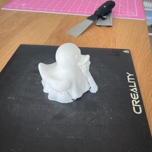 May include: A white 3D-printed duck figurine with detailed feather and wing textures. The duck is positioned on a black surface with the word "CREALITY" printed on the side. A metal spatula and pink cutting mat are in the background.