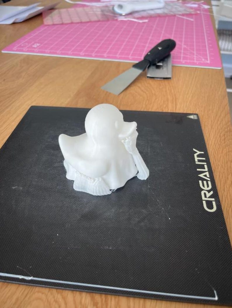 May include: A white 3D-printed duck figurine with detailed feather and wing textures. The duck is positioned on a black surface with the word "CREALITY" printed on the side. A metal spatula and pink cutting mat are in the background.
