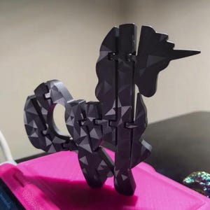 Puede incluir: Un rompecabezas de unicornio geométrico negro con una superficie texturizada. El unicornio está ensamblado a partir de varias piezas entrelazadas, creando un efecto 3D. El rompecabezas está sobre un fondo rosa.