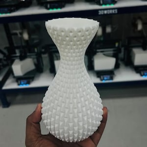 Op de afbeelding: Een witte, getextureerde vaas met een uniek geometrisch patroon. De vaas heeft een brede basis, een smal middengedeelte en een uitlopende bovenkant. Het oppervlak is bedekt met kleine, verhoogde driehoekige vormen. De vaas wordt in de hand van een persoon gehouden.