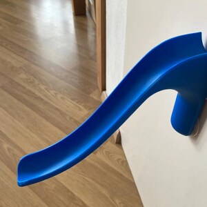 May include: A bright blue plastic slide toy, designed for indoor use. The slide is mounted on a white wall with suction cups. The curved design is reminiscent of a miniature water slide. The background features a wooden floor.