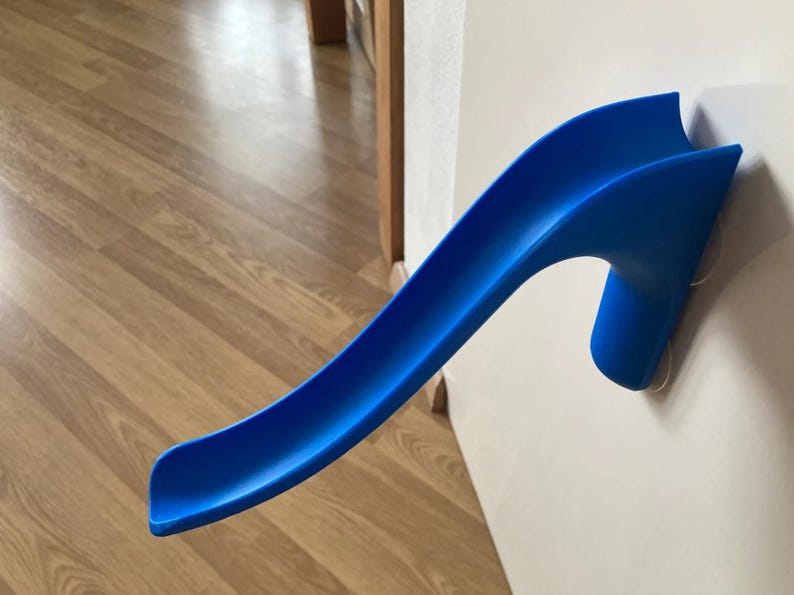 May include: A bright blue plastic slide toy, designed for indoor use. The slide is mounted on a white wall with suction cups. The curved design is reminiscent of a miniature water slide. The background features a wooden floor.