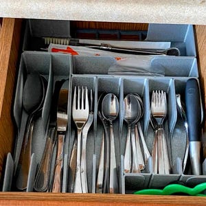 Könnte beinhalten: Eine offene Holzschublade mit einem grauen Besteck-Organizer. Die Schublade enthält verschiedene Besteckteile, darunter Löffel, Gabeln und Messer. Eine Zahnbürste und Zahnpasta sind ebenfalls sichtbar. Das Besteck ist ordentlich angeordnet.